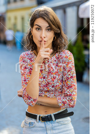 Young woman presses finger to lips makes silence hush sign do not tells gossip secret quiet in city Young woman presses finger to lips makes silence hush sign do not tells gossip secret quiet in city 119250072
