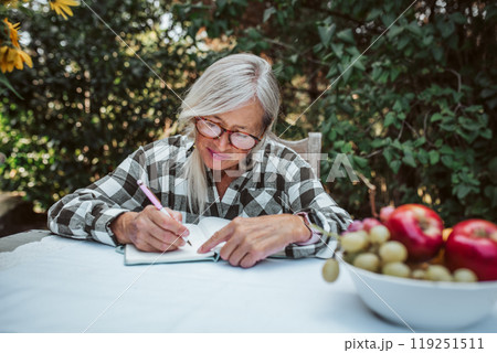 Beautiful older woman is sitting in the garden and writing in her diary. Female senior is keeping daily journal, writing down thoughts and feelings. Beautiful older woman is sitting in the garden and writing in her diary. Female senior is keeping daily journal, writing down thoughts and feelings. 119251511