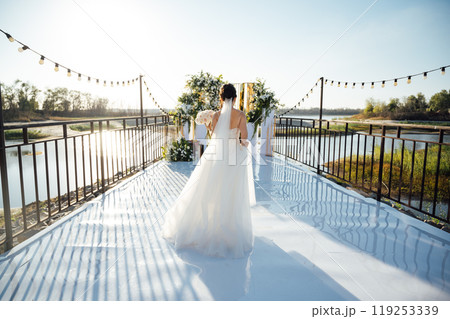 A brunette bride in a white dress holds a bouquet and walks to the wedding arch, among many white chairs. Wedding ceremony in nature. Celebration 119253339
