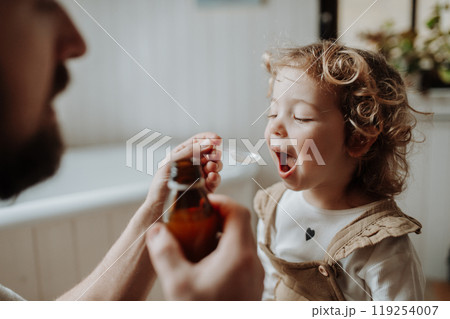 Sick girl with fever and cold at home. Father giving his daughter medicine on spoon, cough syrup or fever medicine. Sick girl with fever and cold at home. Father giving his daughter medicine on spoon, cough syrup or fever medicine. 119254007