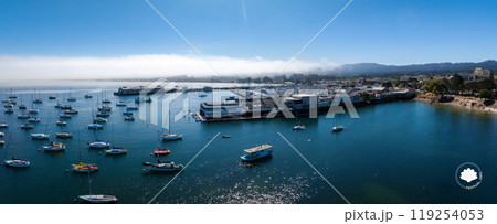 An aerial view captures Monterey, California's harbor filled with sailboats and yachts. Waterfront buildings line the shore, with fog over distant hills. An aerial view captures Monterey, California's harbor filled with sailboats and yachts. Waterfront buildings line the shore, with fog over distant hills. 119254053