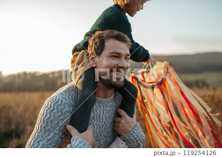 Rear view of father walking with daughter on his shoulders across autumn meadow. Girl holding wreath with ribbons. 119254126