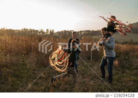 Portrait of young family flying a kite in the autumn nature, jumping in the middle meadow during sunset. Indian summer. 119254128
