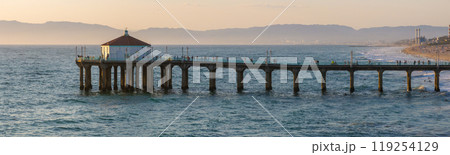 The Manhattan Beach Pier extends into the Pacific Ocean, featuring a roundhouse. Gentle waves meet the sandy shore, with hills and a clear sky in the background. 119254129