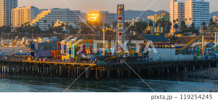 The Santa Monica Pier extends into the Pacific Ocean, featuring a Ferris wheel and roller coaster. High rise buildings in the background catch the sunset light. 119254245
