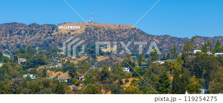 The Hollywood Sign stands prominently on Los Angeles hills, surrounded by rugged terrain, scattered vegetation, and residential homes under a clear sky. 119254275
