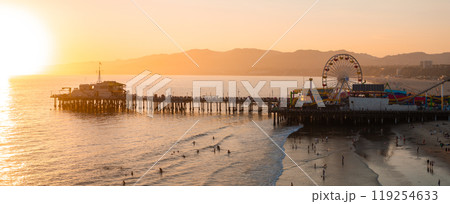 The Santa Monica Pier extends into the Pacific Ocean at sunset, featuring the illuminated Ferris wheel. People enjoy the beach with distant mountains visible. 119254633