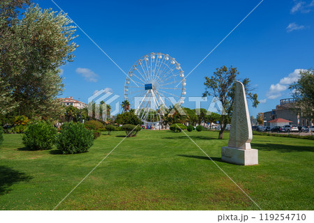 A lively park in Cagliari, Sardinia, featuring a large Ferris wheel, lush greenery, and a white stone monument under a clear blue sky. 119254710