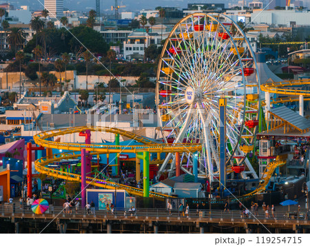 The image captures Santa Monica Pier's Pacific Park, highlighting the Ferris wheel and roller coaster. Visitors explore the attractions with the cityscape behind. 119254715
