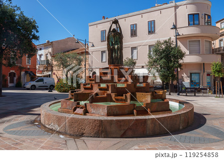 A tiered stone fountain with a bronze statue in a Sardinian square, surrounded by Mediterranean buildings and trees under a clear blue sky. 119254858
