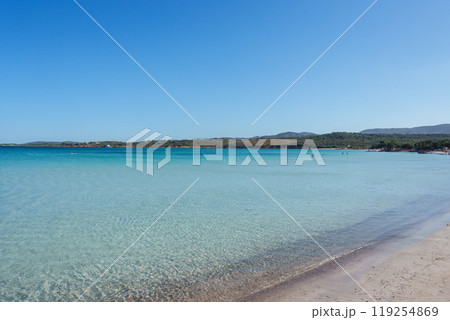 Experience tranquility on a pristine Sardinian beach with crystal clear waters, sandy shores, and a picturesque backdrop of hills under a bright blue sky. 119254869