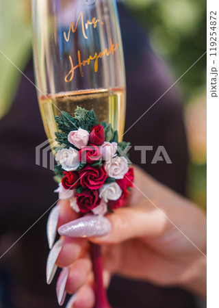 A hand with glittery nails holds a champagne flute personalized with 'Mrs. Hayak'. The stem is adorned with red and white roses, set against a blurred background. A hand with glittery nails holds a champagne flute personalized with 'Mrs. Hayak'. The stem is adorned with red and white roses, set against a blurred background. 119254872