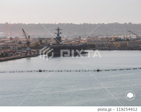 An aerial perspective captures a naval aircraft carrier at San Diego harbor, surrounded by industrial buildings, cranes, and a hilly landscape. An aerial perspective captures a naval aircraft carrier at San Diego harbor, surrounded by industrial buildings, cranes, and a hilly landscape. 119254912
