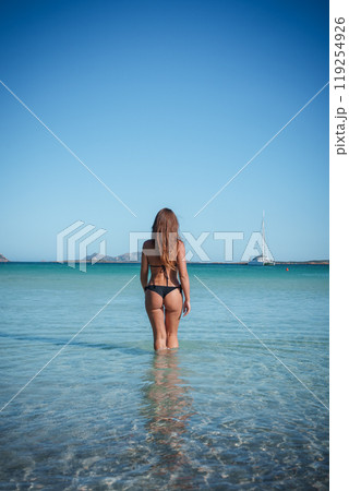 A serene scene in Sardinia featuring a woman standing in clear waters, with a white sailboat anchored in the distance under a clear blue sky. A serene scene in Sardinia featuring a woman standing in clear waters, with a white sailboat anchored in the distance under a clear blue sky. 119254926