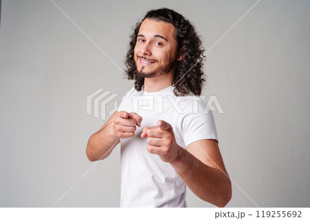 A smiling young man with curly hair playfully pointing directly at the viewer in a light-colored setting A smiling young man with curly hair playfully pointing directly at the viewer in a light-colored setting 119255692
