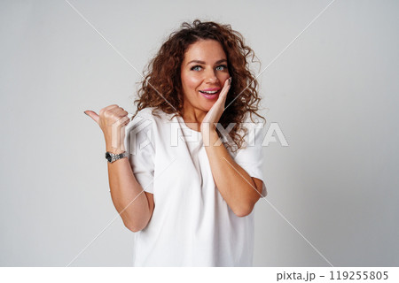 A young woman with curly hair playfully pointing finger while standing against a gray background 119255805