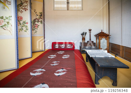 Traditional Korean bedroom at Namsangol Hanok Village featuring a red bedspread with crane motifs, a wooden side table, and floral folding screens. The serene decor highlights the cultural heritage. Traditional Korean bedroom at Namsangol Hanok Village featuring a red bedspread with crane motifs, a wooden side table, and floral folding screens. The serene decor highlights the cultural heritage. 119256348