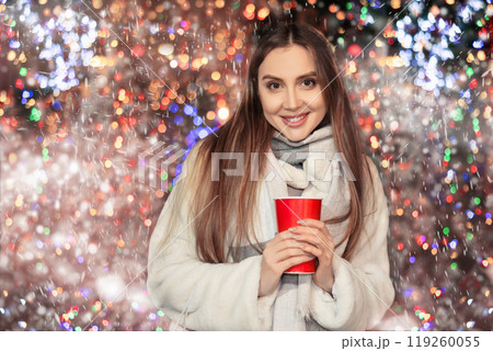 Girl in fur coat stands with glass of coffee in hands on shiny flashing festive sparkling background Girl in fur coat stands with glass of coffee in hands on shiny flashing festive sparkling background 119260055