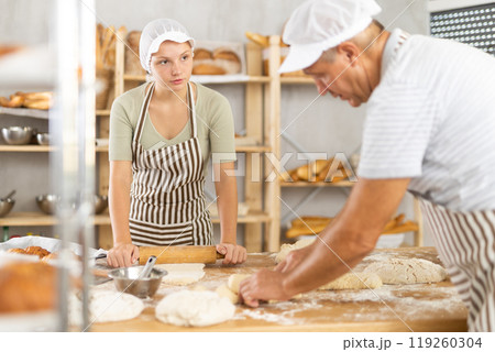 Elderly baker teaches young girl how to make raw dough - rolling it out with rolling pin 119260304