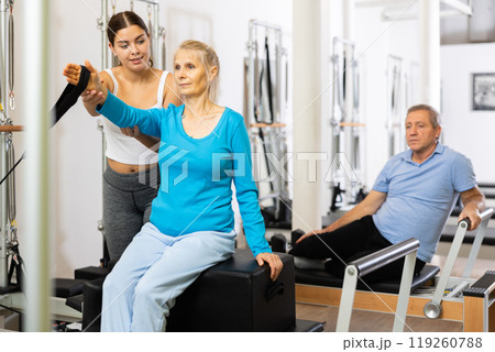 Experienced personal female trainer controlling movements of mature woman exercising on Pilates reformer bed in studio Experienced personal female trainer controlling movements of mature woman exercising on Pilates reformer bed in studio 119260788