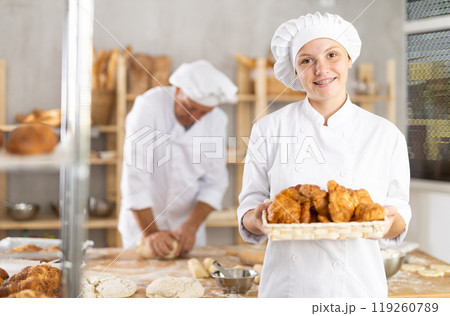Smiling young girl baker in uniform stands behind counter in small private bakery offering croissants 119260789