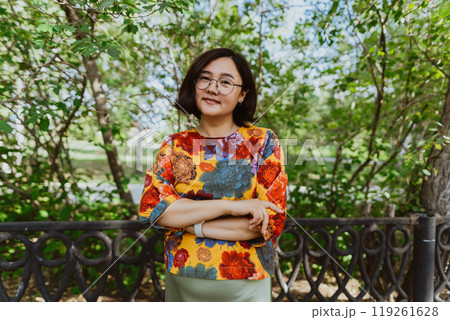 Smiling female with crossed arms standing in a lush park, exuding happiness Confident asian woman in glasses and bright blouse with gorgeous smile posing in summer park. Portrait looking at camera Smiling female with crossed arms standing in a lush park, exuding happiness Confident asian woman in glasses and bright blouse with gorgeous smile posing in summer park. Portrait looking at camera 119261628