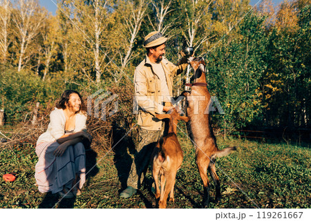 A man farmer with a beard and a hat on a small farm feeds Nubian goats on a sunny, peaceful day. Scene showcases the connection between the farmer and her animals, emphasizing eco-friendly farming A man farmer with a beard and a hat on a small farm feeds Nubian goats on a sunny, peaceful day. Scene showcases the connection between the farmer and her animals, emphasizing eco-friendly farming 119261667