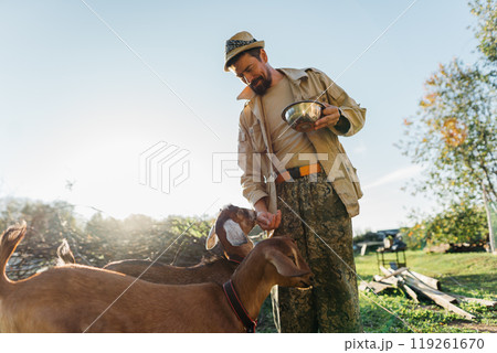 A man farmer in a hat feeds Nubian goats oats from a bowl on his free-range farm. Nubian goats being fed oats by a man on her organic farm A man farmer in a hat feeds Nubian goats oats from a bowl on his free-range farm. Nubian goats being fed oats by a man on her organic farm 119261670
