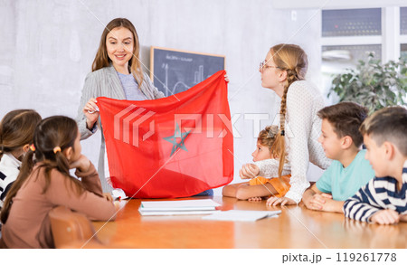 Smiling young female teacher conducting lesson for preteens, showing state flag of Morocco in audience of middle school 119261778