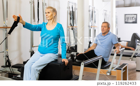 Mature woman in sportswear sitting and doing Pilates exercises for hands on reformer bed equipment in fitness center Mature woman in sportswear sitting and doing Pilates exercises for hands on reformer bed equipment in fitness center 119262433