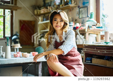 Pretty female artist smiling and posing at creative workshop with her artwork in the background 119265982
