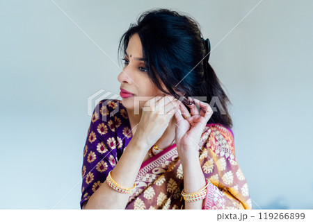 Dressed in beautiful saree, Indian woman delicately putting traditional jewellery getting ready for an ethnic festive celebration. Indian culture and ritual holidays in modern life. Selective focus. 119266899