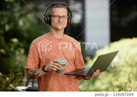 Medium portrait shot of smiling businessman in headphones holding laptop and reusable cup captured during outdoor freelance work in park, copy space Medium portrait shot of smiling businessman in headphones holding laptop and reusable cup captured during outdoor freelance work in park, copy space 119267264
