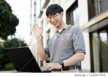 Medium shot of smiling Asian businessman waving hand looking at laptop screen captured during online conference while working at stand up desk outside in city street, copy space 119267304