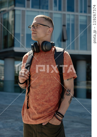 Vertical shot of tattooed man wearing glasses, backpack and casual outfit posing outside in the sunlight keeping one hand in pocket in city street, copy space 119267374