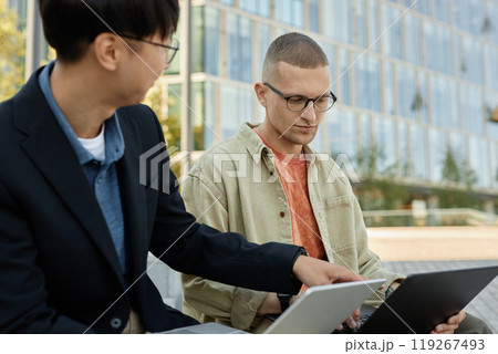 Medium shot of stylish man with buzz cut and glasses listening to coworker while collaborating on work looking at laptop screen during outdoor meeting, copy space Medium shot of stylish man with buzz cut and glasses listening to coworker while collaborating on work looking at laptop screen during outdoor meeting, copy space 119267493