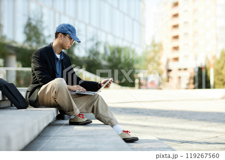 Side view of Asian male IT programmer writing code on laptop looking at computer screen while sitting on concrete steps in city street, copy space Side view of Asian male IT programmer writing code on laptop looking at computer screen while sitting on concrete steps in city street, copy space 119267560