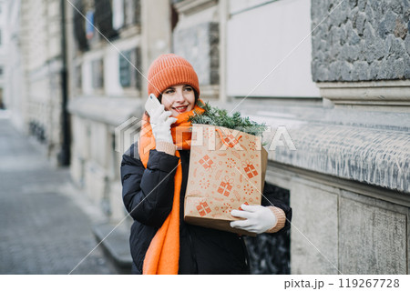 Preparing for Christmas. Woman in orange hat and scarf holding festive shopping bag while speaking on the phone, winter holiday shopping concept Preparing for Christmas. Woman in orange hat and scarf holding festive shopping bag while speaking on the phone, winter holiday shopping concept 119267728