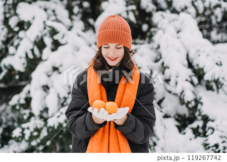 Woman in orange winter attire holding a tangerine, smiling playfully in snow-covered surroundings. Winter wellness, mindfulness, and seasonal joy. 119267742