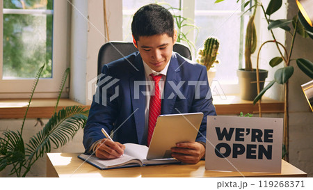 Businessman in formal wear sitting at desk with we are open sign and signing documents, checking information with tablet. 119268371