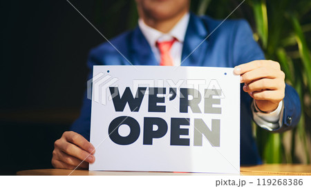 Cropped image of man in formal wear sitting in with we are open sign in hands, meaning opening new business, promotion of new ventures. Cropped image of man in formal wear sitting in with we are open sign in hands, meaning opening new business, promotion of new ventures. 119268386