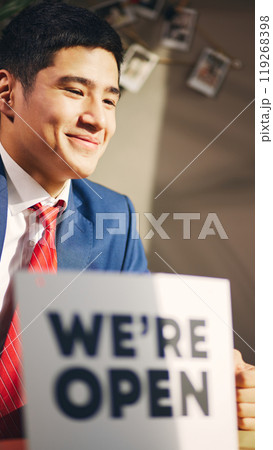 Smiling young man and formal wear sitting in sunlight with we are open sign, meaning opening new business. Smiling young man and formal wear sitting in sunlight with we are open sign, meaning opening new business. 119268398