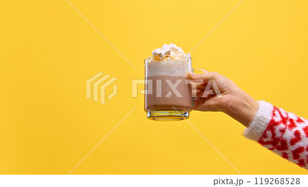 Hand in cozy sweater holds glass mug of hot chocolate topped with fluffy marshmallows against warm yellow studio background. Hand in cozy sweater holds glass mug of hot chocolate topped with fluffy marshmallows against warm yellow studio background. 119268528