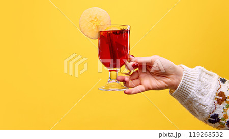 Woman's hand holds a glass of mulled wine, garnished with apple slice and cinnamon stick, against warm yellow studio background. Mulled wine. Woman's hand holds a glass of mulled wine, garnished with apple slice and cinnamon stick, against warm yellow studio background. Mulled wine. 119268532