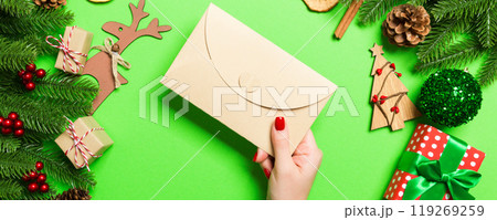 Top view of woman holding an envelope on green background made of holiday decorations. Christmas time concept Top view of woman holding an envelope on green background made of holiday decorations. Christmas time concept 119269259