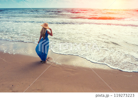 Seascape during sunrise. Woman on the beach. Young happy woman in a long fluttering blue dress and straw hat walks on the seashore 119273223