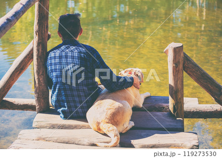Man with Labrador retriever dog sitting on a wooden deck by the lakeshore and looking at water 119273530