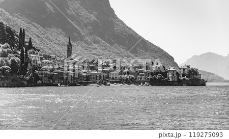 The picturesque town of Menaggio hugs the shores of Lake Como, showcasing houses, a bell tower, and greenery under the warm sun. Black and white image. The picturesque town of Menaggio hugs the shores of Lake Como, showcasing houses, a bell tower, and greenery under the warm sun. Black and white image. 119275093