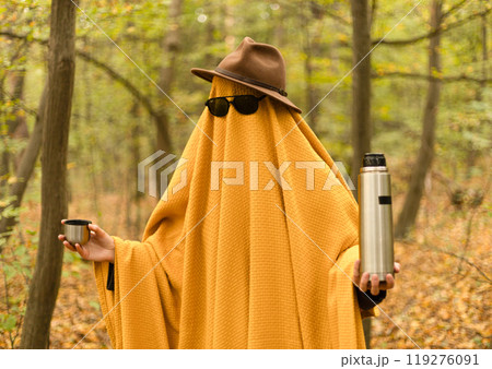 ghost in a yellow blanket, hat and glasses in the autumn forest with a thermos 119276091