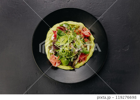 Fresh salad with avocado cream and prosciutto, served on a black plate, perfect for gourmet dining 119276698